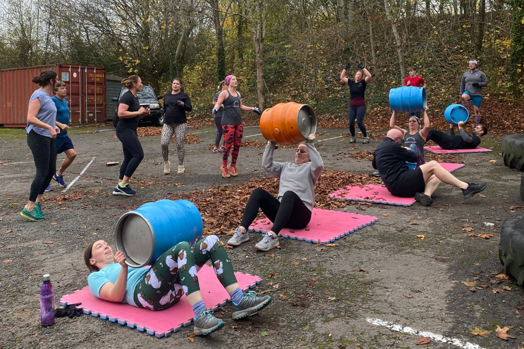 A photograph showing an outdoor group exercise bootcamp. People are lifting barrels over their heads.
